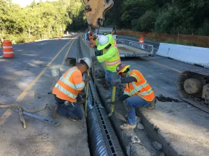 Construction crew installing corrugated steel slotted drain for roadway drainage