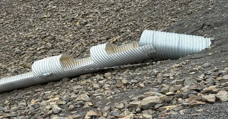 Close view of modified corrugated steel downdrain sections with open half-round channels and full-round connection points on a rocky Idaho highway slope.