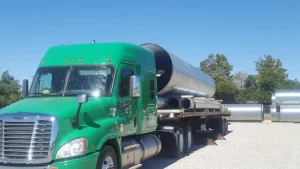 Green semi-truck transporting corrugated steel pipe on a flatbed trailer.