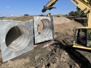 Burke SD Triple 120in CHS (4)_resized Excavator positioning a prefabricated steel culvert headwall during the County Road 27 replacement project in South Dakota.